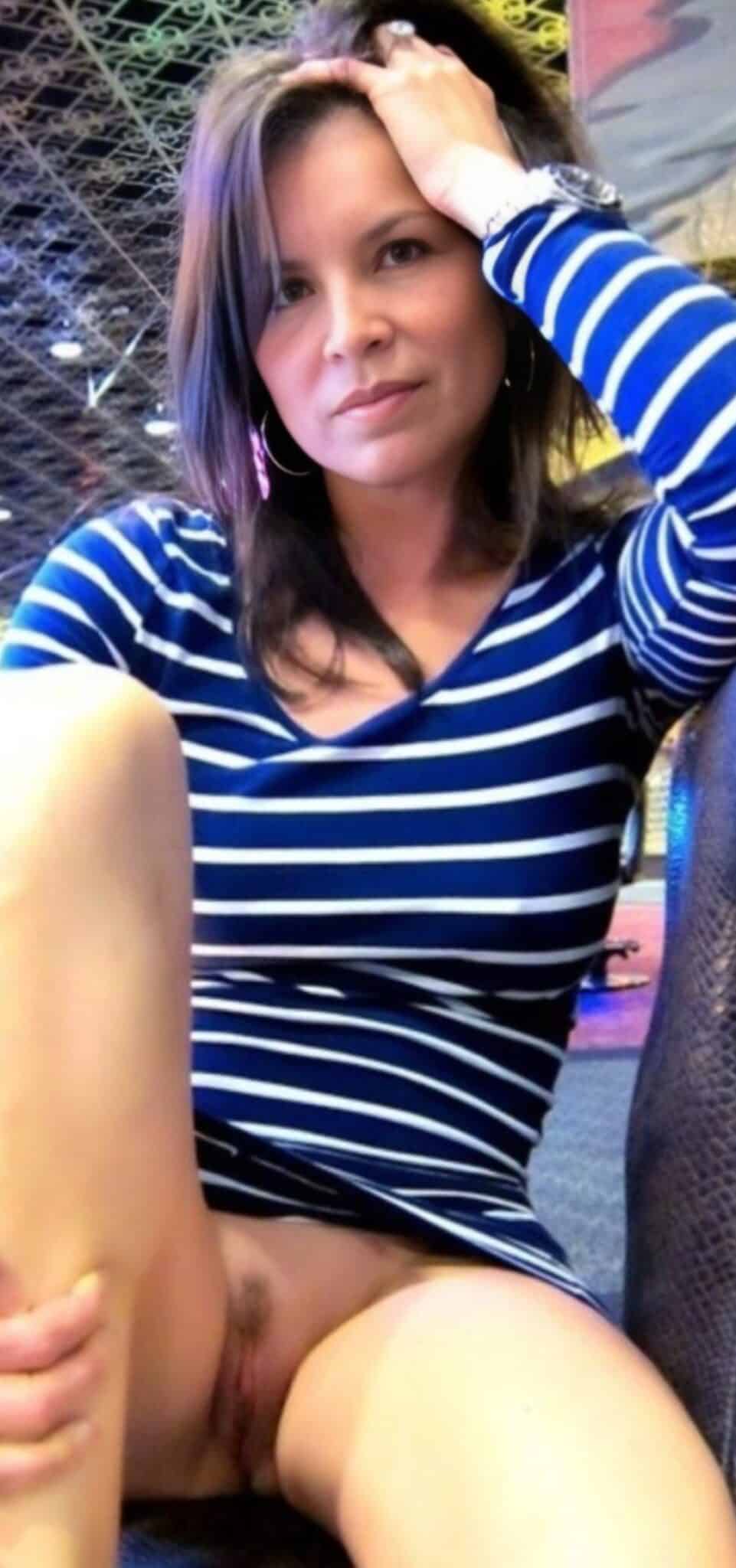 Amateur: What’s the Deal with This Latina’s No-Panties Pose? A Latina woman poses confidently in a bedroom, legs spread, no panties, wearing a striped top, hoop earrings, and a watch.